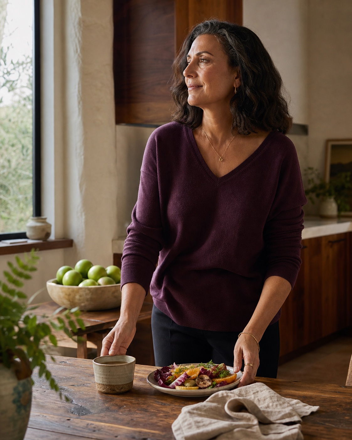 A woman in a plum sweater sets a colorful plate beside tea in a sunlit kitchen, with a bowl of green apples nearby